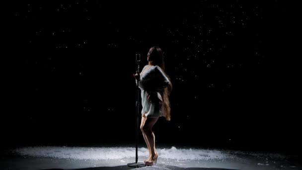 Belle jeune femme dans une robe brillante du soir chante dans un microphone vintage sur fond de neige tombante. Le soliste sourit et se déplace élégamment sur un fond noir. Mouvement lent. — Video