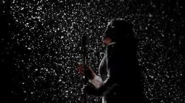 Güzel bir şarkıcının siluetleri ve kar yağan arka planda klasik bir mikrofonu var. Stüdyo ışığında kar taneleri parlar. Yan görüş. Ağır çekim. Kapat..