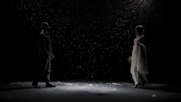 Happy newlywed couple approach each other against the background of falling snow. A man in a suit and a woman in a white long dress with a bouquet on a black background. Slow motion. — Stock Video