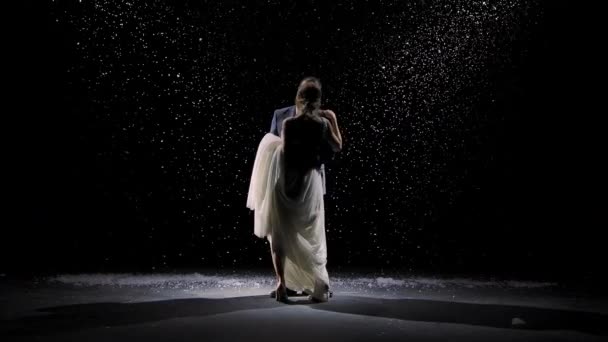 Una encantadora pareja de enamorados baila sobre el fondo de la nieve que cae. Un hombre con un traje y una mujer con un vestido largo blanco sobre un fondo negro en los rayos de luz del estudio. Movimiento lento. — Vídeo de stock