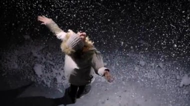 Şapkalı ve kalın elbiseli mutlu genç bir kadının yağan karlar arasında dönüşünün en üst görüntüsü. Büyük kar taneleri siyah arka plana karşı ağır çekimde havada yüzer. İlk kar yağışı. Kapat..