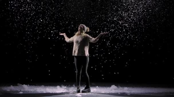 Gelukkige jonge vrouw met een hoed en een warme trui draait tussen de vallende sneeuw. Grote sneeuwvlokken zweven in slow motion tegen een zwarte achtergrond in de lucht. Nieuwjaar of Kerst schieten. — Stockvideo