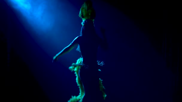 A dark silhouette of an attractive woman in a revealing carnival costume and a headdress with feathers passionately dances on a black background with blue light. Close up. — Stock Video