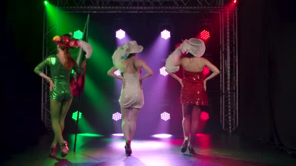 Tres elegantes bailarines de cabaret con vestidos cortos y brillantes actúan en un estudio oscuro con un telón de fondo de luces de colores brillantes. Una mujer sostiene la bandera de Italia. Movimiento lento. — Vídeos de Stock
