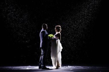 Kar yağan arka planda duran mutlu yeni evli çift. Takım elbiseli bir adam ve beyaz uzun elbiseli bir kadın stüdyo ışıklarının altında siyah bir arka planda buketi olan bir kadın..