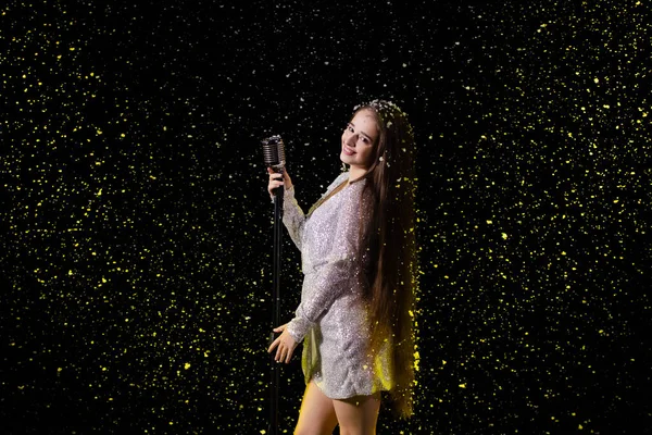 Retrato de una hermosa cantante con un micrófono vintage sobre el fondo de la nieve que cae. Mujer joven en un escenario vacío en los rayos de luz amarilla estudio de cerca. —  Fotos de Stock