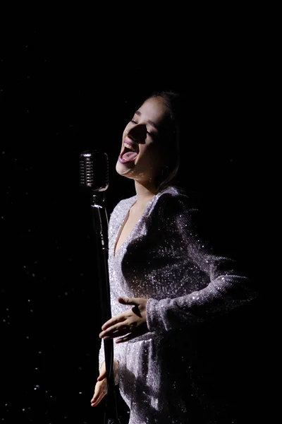 Beautiful young woman in an evening dress sings into a vintage microphone against the background of falling snow in a dark studio. Close up. — Stock Photo, Image