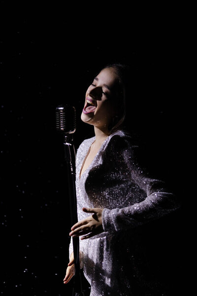 Beautiful young woman in an evening dress sings into a vintage microphone against the background of falling snow in a dark studio. Close up.