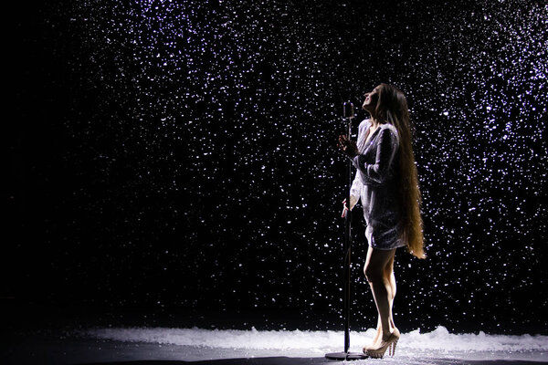 Side view of a charming young woman in an evening shiny dress singing into a vintage microphone against the background of falling snow.