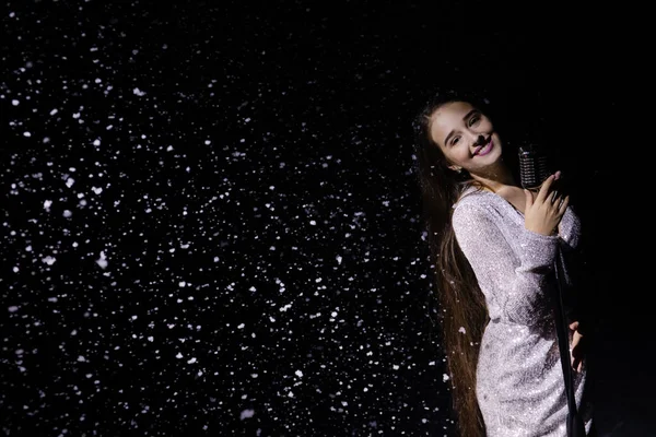 在降雪的背景下,美丽的歌手拿着老式话筒摆姿势.雪花在演播室的灯光下闪闪发光.靠近点. — 图库照片