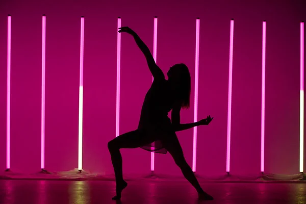 Silhouette di una giovane ballerina che balla e posa in una stanza con lampade al neon. Un modello snello e moderno in una bella posa su uno sfondo viola. — Foto Stock