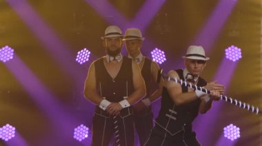 Siyah beyaz takım elbiseli ve şapkalı üç jimnastikçiden oluşan bir grup parlayan sazlıklarla gösteri yapıp onları döndürüyor. Erkekler karanlık bir stüdyoda arka planda mor spot ışıklarına karşı poz verirler. Ağır çekim. Kapat..