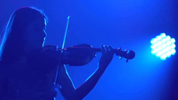 Silhueta de uma bela morena músico feminino em um vestido brilhante toca violino em um estúdio escuro com iluminação azul. Fechar. — Vídeo de Stock