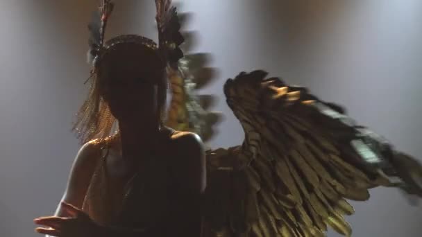 En teaterföreställning med den grekiska gudinnan Artemis. Charmig kvinna i gyllene kostym med vingar dansar och sjunger i en mörk studio med bakgrundsbelysning. Silhuett. Närbild. — Stockvideo