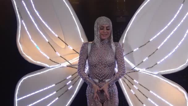 Penyanyi yang ramping dan chic mengenakan jumpsuit ketat yang diisi dengan rhinestones, dengan sayap kupu-kupu bersinar di belakangnya. Dekat dari seorang wanita muda bernyanyi di studio gelap. — Stok Video