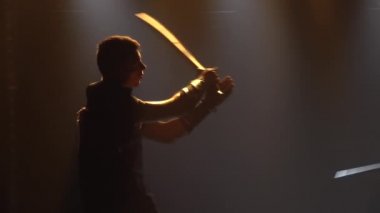 Erkekler karanlık bir stüdyoda kılıçla sahne ışıklarıyla kavga ederler. Zırhlı erkek aktörlerin siluetleri bir tiyatro gösterisinde oynuyorlar. Ağır çekimde kapat..