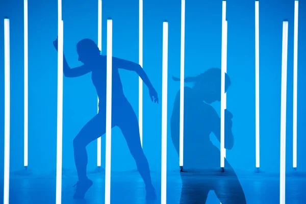 Koláž stínů půvabná mladá žena tančící prvky moderního tance v ateliéru na modrém pozadí s neonovou osvětlovací lampou. Design plakátu tance. — Stock fotografie