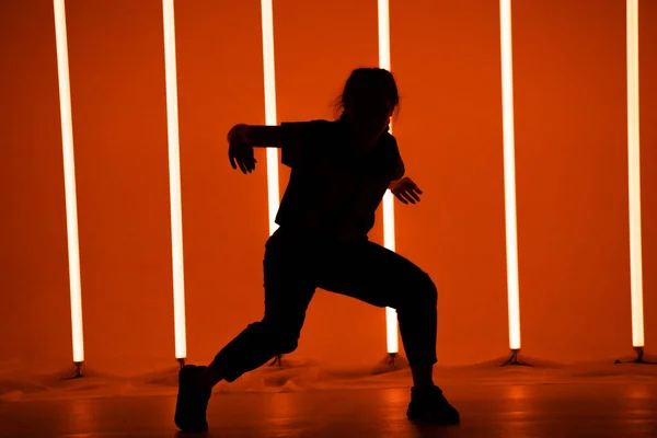 Mladá žena oblečená v pouliční módní oblečení tanec hip-hop styl ve studiu s neonové osvětlení trubice na oranžovém pozadí. Taneční a hudební design plakátů. — Stock fotografie