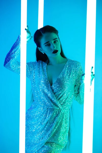 A seductive brunette with long hair in a short shiny dress looks passionately into the camera. Shot against the background of blue neon tubes in the studio. Close up. — Stock Photo, Image