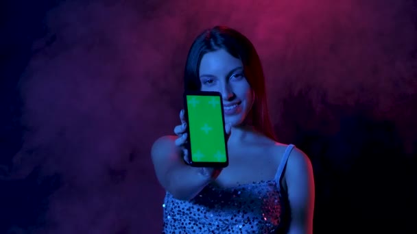 Портрет чарівної молодої жінки дивиться на камеру, показує смартфон із зеленим екраном і вказує на нього пальцем. Близько. Повільний рух . — стокове відео