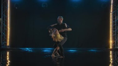 Dans eden güzel çift Latin dansı. Siyah elbiseli profesyonel dansçılar suyun yüzeyinde salsa yapıyor. Arka plandaki karanlık stüdyo. Yavaş çekim.