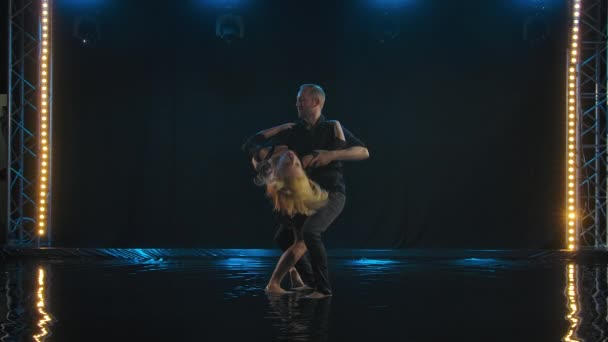 Hermosa pareja bailando baile latino. Bailarinas profesionales en ropa negra bailan salsa en la superficie del agua. Estudio oscuro retroiluminado. Movimiento lento. — Vídeos de Stock