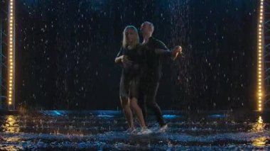 Aktif mutlu bir adam ve bir kadın yağmurda karanlık bir stüdyoda salsa yapıyor. Siyahlı bir çift yağmur damlaları ve su damlaları arasında dans etmekten zevk alıyor. Yavaş çekim.