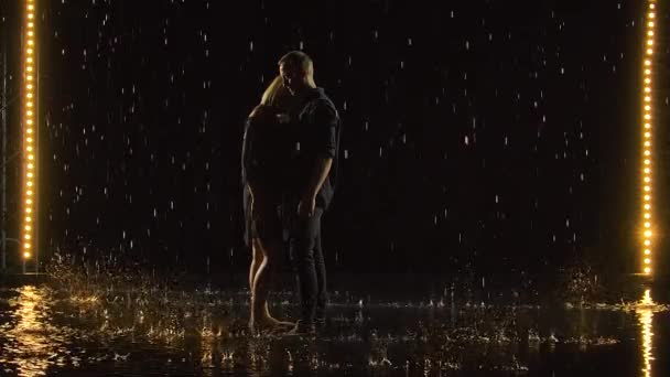魅力的な若いカップルが雨の中で受け入れられ、瞬間を楽しんでいます。雨滴と水しぶきの間に黒いスタジオの背景に男と女。スローモーション. — ストック動画
