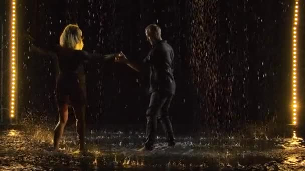 Hombre feliz activo y una mujer bailando salsa juntos en un estudio oscuro bajo la lluvia. Una pareja de negro está disfrutando del baile entre gotas de lluvia y salpicaduras de agua. Movimiento lento. — Vídeos de Stock