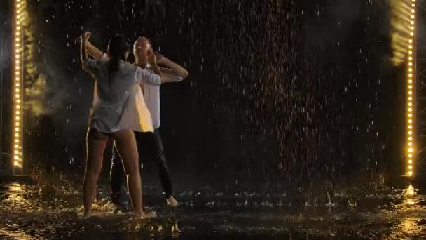 情熱的なぬれたカップルのキスと雨の中で踊る。雨滴の間の愛の物語。ゆっくりとした動きで撮影された黒のスタジオの背景にステージライト. — ストック動画