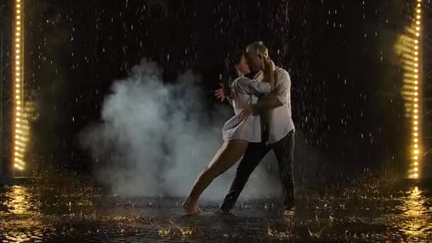 情熱的なぬれたカップルのキスと雨の中で踊る。雨滴の間の愛の物語。ゆっくりとした動きで撮影された黒のスタジオの背景にステージライト. — ストック動画