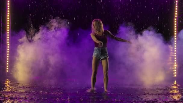 Una encantadora rubia esbelta está bailando salsa en un estudio oscuro entre las gotas de lluvia y las salpicaduras de agua. Joven bailarina se mueve en cámara lenta sobre un fondo de estudio ahumado con retroiluminación púrpura. — Vídeo de stock
