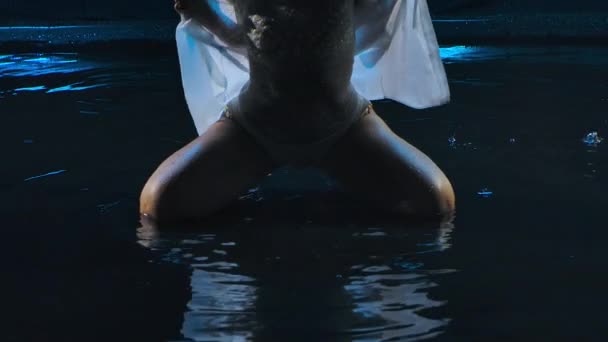 Sexy horká brunetka v bílém obleku vytváří cákance vlasů a pohybuje eroticky ve zpomaleném filmu. Silueta ženy sedící na hladině vody zblízka. — Stock video