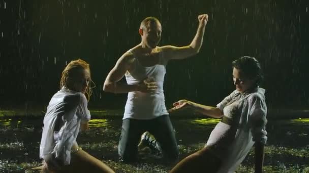 Dos hermosas mujeres jóvenes con camisas blancas mojadas y un hombre bailando apasionadamente bajo la lluvia. Siluetas de cuerpos mojados en el humo. En cámara lenta. De cerca.. — Vídeos de Stock