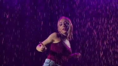 Şortlu ince bir sarışın ve tişörtlü yağmurda salsa dansının tadını çıkarıyor. Tutkulu genç bir kadın ağır çekimde stüdyoda mor ışık ve dumanın arka planına karşı hareket ediyor. Kapat.
