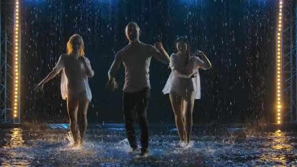 两个女人和一个男人在黑暗的工作室背景下，在雨滴中跳着热情的萨尔萨舞。专业舞蹈演员在水面上一个烟雾弥漫的演播室里练习拉丁舞。慢动作. — 图库视频影像