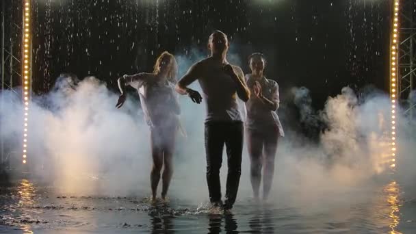 Dos mujeres y un hombre bailando un apasionado baile de salsa en gotas de lluvia sobre un fondo oscuro estudio. Bailarines profesionales practican latino en un estudio ahumado en la superficie del agua. Movimiento lento. — Vídeo de stock