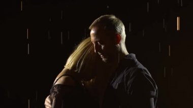 Yağmurda sarılan ve anın tadını çıkaran genç bir çift. Parlak yağmur damlalarının arasında, siyah bir stüdyonun arka planında kadın ve erkeği kapatın. Yavaş çekim.