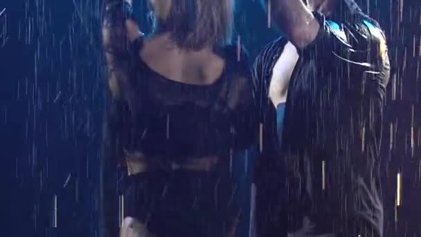 검은 옷을 입은 젖은 한 남자와 한 여자가 빗방울 사이에서 빙빙 돌면서 열정적으로 춤을 추고 있다. 한 쌍의 커플이 어두운 스튜디오에서 무대 조명과 함께 춤을 추고 있습니다. 슬로우 모션으로 닫기. — 비디오