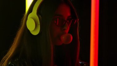 Güzel genç bir kadının portresi. Büyük beyaz kulaklıklarla müzik dinliyor ve sakızdan baloncuklar üflüyor. Kapatın. Yavaş çekim.