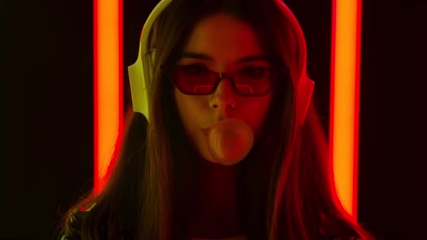 Portrait of a pretty young beautiful woman listening to music in big white headphones and blowing bubbles from gum. Close up. Slow motion. — Stock Video