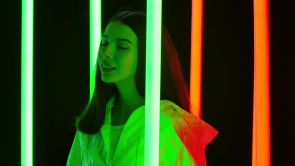 Portret van een mooie jonge mooie vrouw die poseert tegen een donkere studio achtergrond met felgekleurde neon buizen. Sluit maar af. Langzame beweging. — Stockvideo