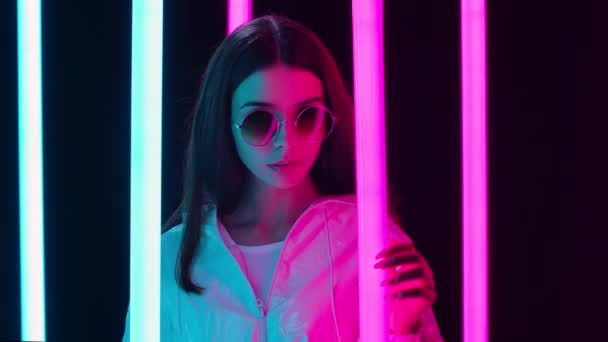 Retrato de uma bela mulher muito jovem em óculos de sol redondos elegantes posando contra um fundo de estúdio escuro com tubos de néon multicoloridos brilhantes. Fecha. Movimento lento. — Vídeo de Stock