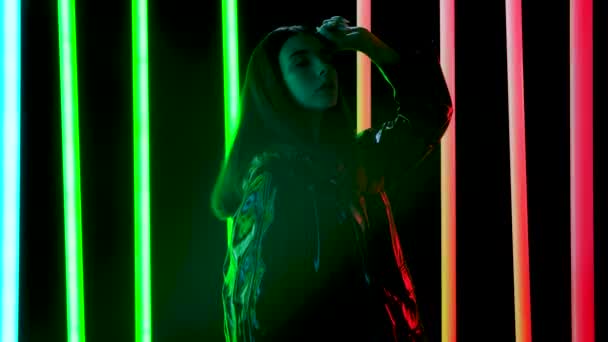 Retrato de una joven mujer bonita posa sobre un fondo de estudio oscuro con tubos de neón multicolores brillantes. Movimiento lento. — Vídeo de stock