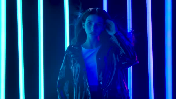 Portrait of a young pretty woman with hair flying in the wind poses against a dark studio background with bright multicolored neon tubes. Slow motion. — Stock Video