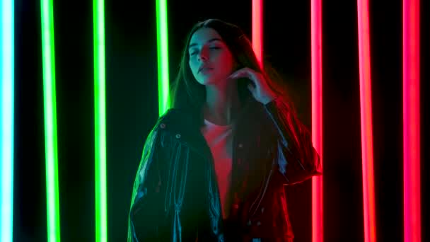 Portrait of a young pretty woman with hair flying in the wind poses against a dark studio background with bright multicolored neon tubes. Slow motion. — Stock Video