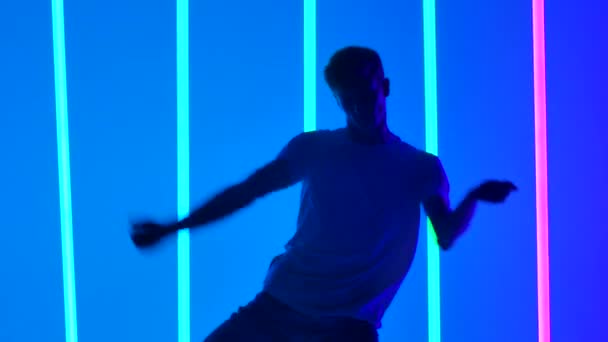 Genç sokak kıyafetleri giymiş, hip hop tarzı gösteri yapan aktif bir genç adamın siluetleri. Bir dansçı erkek, bir alacakaranlık stüdyosunda parlak neon ışıklarının önünde yaratıcılık pratiği yapar. Kapatın. Yavaş çekim — Stok video