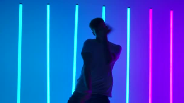 Silhouette di un giovane uomo attivo vestito di streetwear giovanile che si esibisce in stile hip hop. Un ballerino maschio pratica la creatività davanti a luci al neon in uno studio crepuscolare. Chiudete. Rallentatore — Video Stock