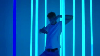 Genç sporcu, parlak mavi neon ışıkların önünde hip hop dansı yapıyor. Sıradan genç kıyafetleri içinde siluetli bir erkek dansçı karmaşık hareketler yapar ve atlar. Kapatın. Yavaş çekim.