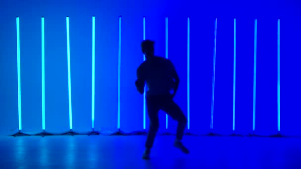 Jonge sportieve man dansend hiphop tegen een achtergrond van felblauwe neon lichten. Een silhouetted mannelijke danser in casual jeugdkleding voert complexe bewegingen en sprongen. Langzame beweging. — Stockvideo